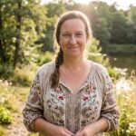 Christine lächelt in die Kamera, im Hintergrund sommerliche Natur mit See im Sonnenlicht